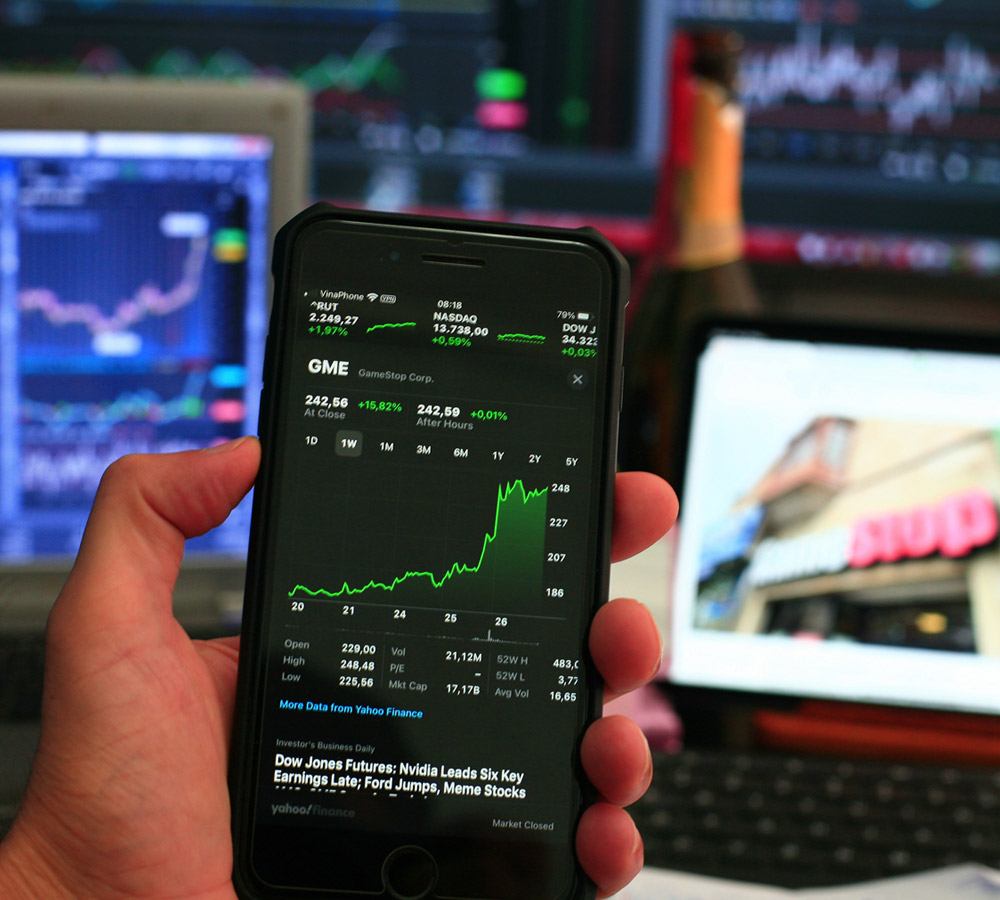 An investor's phone showing a stock chart surging, with trading screens glowing in the background